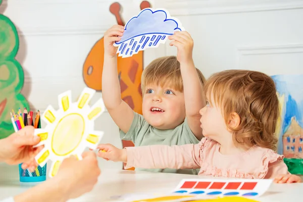 Little cute boy in kindergarten class hold the cart with weather rain card in hand showing to teacher who holds sun image