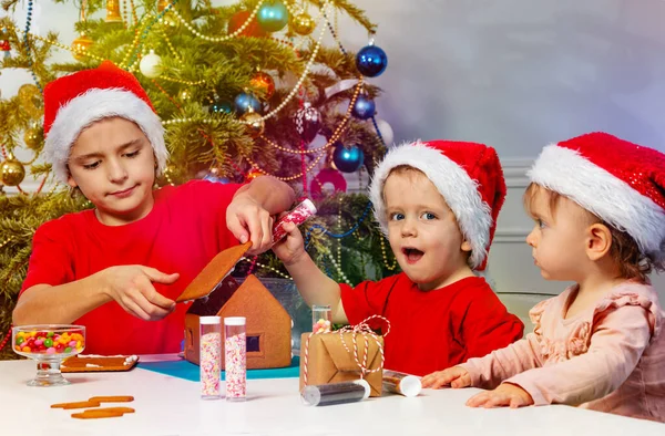 Two Boys Little Girl Santa Hat Gluing Together Gingerbread House — ストック写真