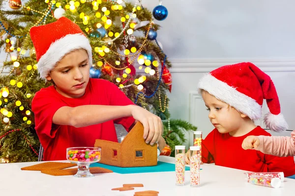 Two Boys Decorate Gingerbread House Sitting Santa Hats Christmas Tree — Zdjęcie stockowe