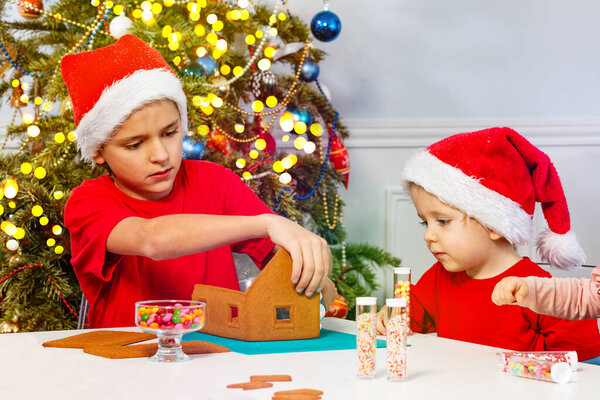 Two boys decorate gingerbread house sitting in Santa hats near Christmas tree