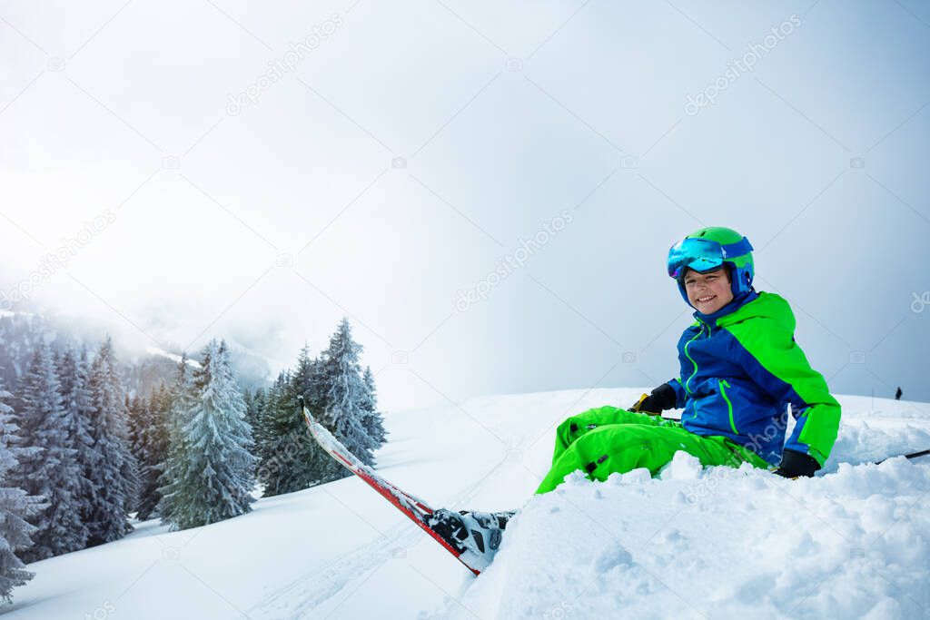 Side view of the boy with alpine ski sit in the snow over snowy forest after strong storm