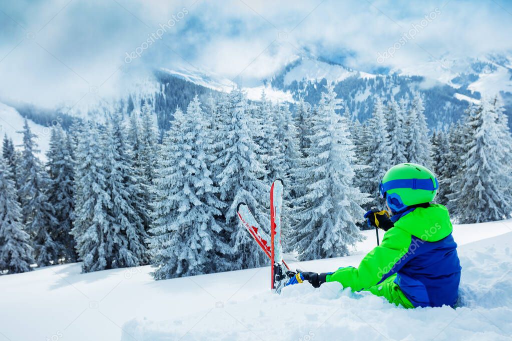 Skier boy sit in the snow over beautiful scenic mountains and snowy fir forest after heavy snowfall view from the back