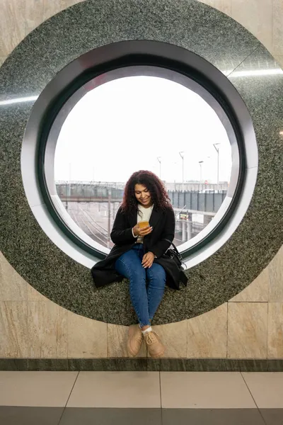 रेलवे स्टेशन पर एक युवा महिला गोल खिड़की पर बैठी है और यो स्मार्टफ़ोन देख रही है रॉयल्टी फ़्री स्टॉक फ़ोटो