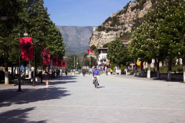 Berat, Arnavutluk - 6 Mayıs 2021. Çiçekli kestaneleri olan yeşil bir meydan, yürüyen insanlar ve bisikletli bir adam. Atmosfer eski Balkan kenti