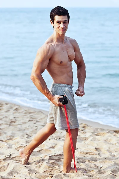 Jeune homme athlétique faisant de l'exercice et de remise en forme avec un extenseur de poitrine (bande de résistance) sur la plage — Photo