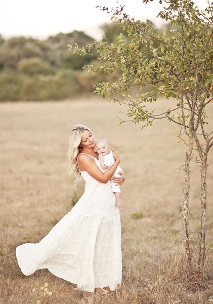 Familia feliz al atardecer. Madre en vestido blanco con bebé en la naturaleza —  Fotos de Stock