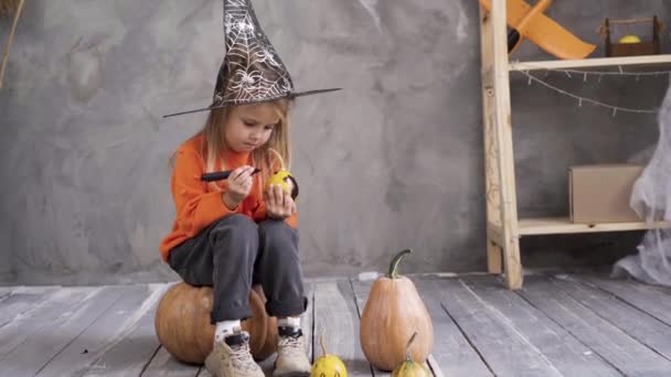 Nahaufnahme eines kleinen kaukasischen Mädchens in der Verkleidung einer Hexe, das auf einem großen Kürbis im Raum in der Verkleidung einer Hexe sitzt und einen Kürbis für das Halloween-Fest bemalt. DIY-Dekor. — Stockvideo