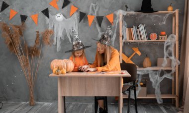 Cadı kostümlü bir kadın ve çocuk evdeki masada oturmuş kendi elleriyle dekor hazırlıyorlar. Cadılar Bayramı için bir oda dekore ediyorum. Anne ve bebeğin birlikteliği.
