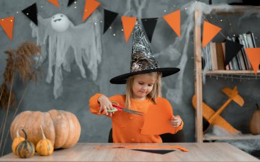 Cadı kostümlü beyaz kız evde masada oturuyor ve Cadılar Bayramı için el sanatları hazırlıyor. Kağıttan yapılmış odada ölüm ve dekor.