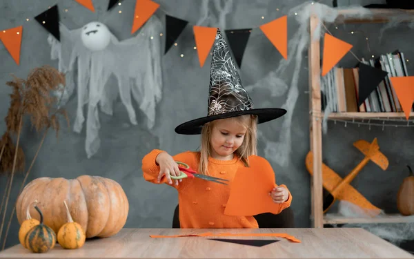 Cadı kostümlü beyaz kız evde masada oturuyor ve Cadılar Bayramı için el sanatları hazırlıyor. Kağıttan yapılmış odada ölüm ve dekor.
