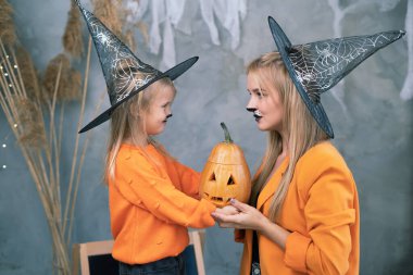 Cadılar Bayramı 'nı balkabağı kabaklı cadı şapkası takmış aile anne ve kızı evde kutluyor. 31 Ekim 'deki Cadılar Bayramı festivalinde makyajlı bir kadın ve bir kız. Gri arkaplan