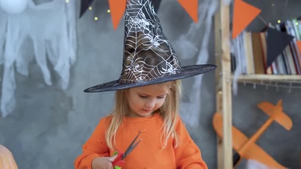 Közelkép egy kislányról egy boszorkány képében, aki a saját kezével vágja ki a papírdekorációt. Készülődés Halloweenre. — Stock videók
