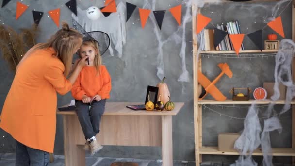 Žena a dítě se připravují na Halloweenské prázdniny. Máma nanáší make-up na dítě ve formě lva doma v místnosti. Čarodějnický klobouk. — Stock video