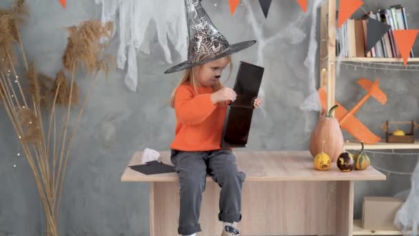 Une petite fille s'assoit à la maison sur la table et regarde le maquillage en l'honneur des vacances d'Halloween. — Video