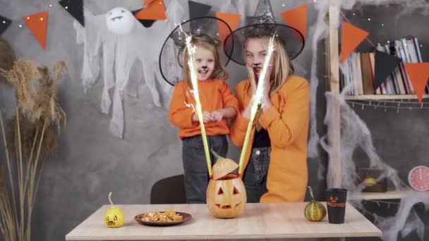 Gelukkig moeder met dochter dragen Halloween hoeden hold blik op oranje pompoen jack-o-lantaarn vuurwerk voor feest in ingerichte kamer. Halloween concept — Stockvideo