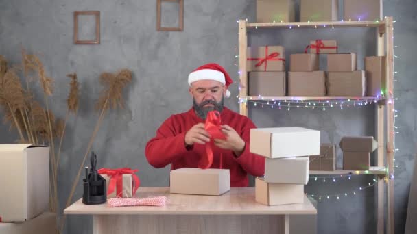 Concept de petite entreprise pour Noël un homme barbu dans un santa claus chapeau assis à une table et colle un grand arc rouge sur une boîte. — Video