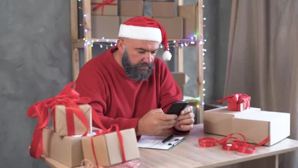 Caucasian male business owner sits at home in the office at the table and takes orders online using his smartphone. Wearing a santa claus hat. Concept for the sale of Christmas goods. — Stock Video
