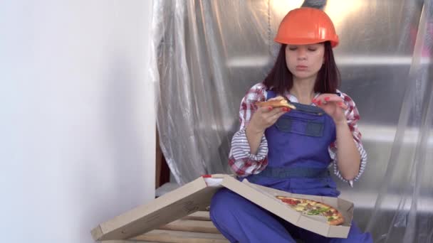 Linda mujer joven constructora con casco trabajando en la renovación de apartamentos. Construcción, reparación y renovación. tomó un descanso y está comiendo pizza. — Vídeo de stock