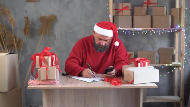 Homem caucasiano com uma barba e um chapéu de Papai Noel senta-se em um escritório em casa e leva uma ordem on-line para produtos usando o telefone. Faz anotações em papel com uma caneta. — Vídeo de Stock