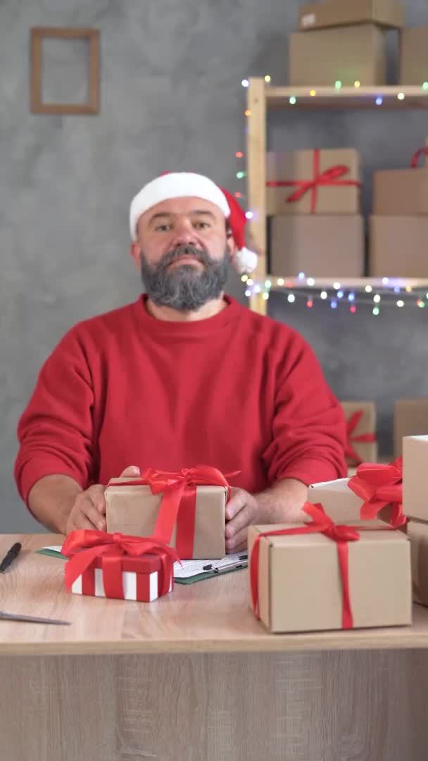Online obchodování. U stolu v kanceláři sedí běloch v klobouku Santa Clause a s bílým vousem a v rukou drží krabici s červeným lukem. Selektivní soustředění. Kopírovat prostor. — Stock video