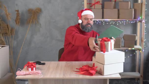 Homem caucasiano com barba e chapéu de Papai Noel prepara mercadorias para embarque no escritório de casa. Ela dobra caixas e faz anotações em papel com uma caneta. Conceito para a venda de Natal ou Ano Novo. — Vídeo de Stock