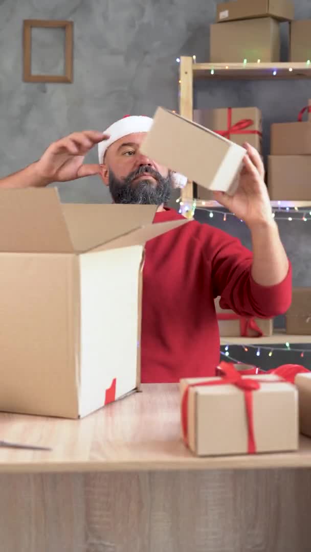 Hombre caucásico en sombrero de santa claus y barba blanca prepara un gran pedido para el envío. Toma cajas pequeñas y las apila en una caja grande. — Vídeos de Stock