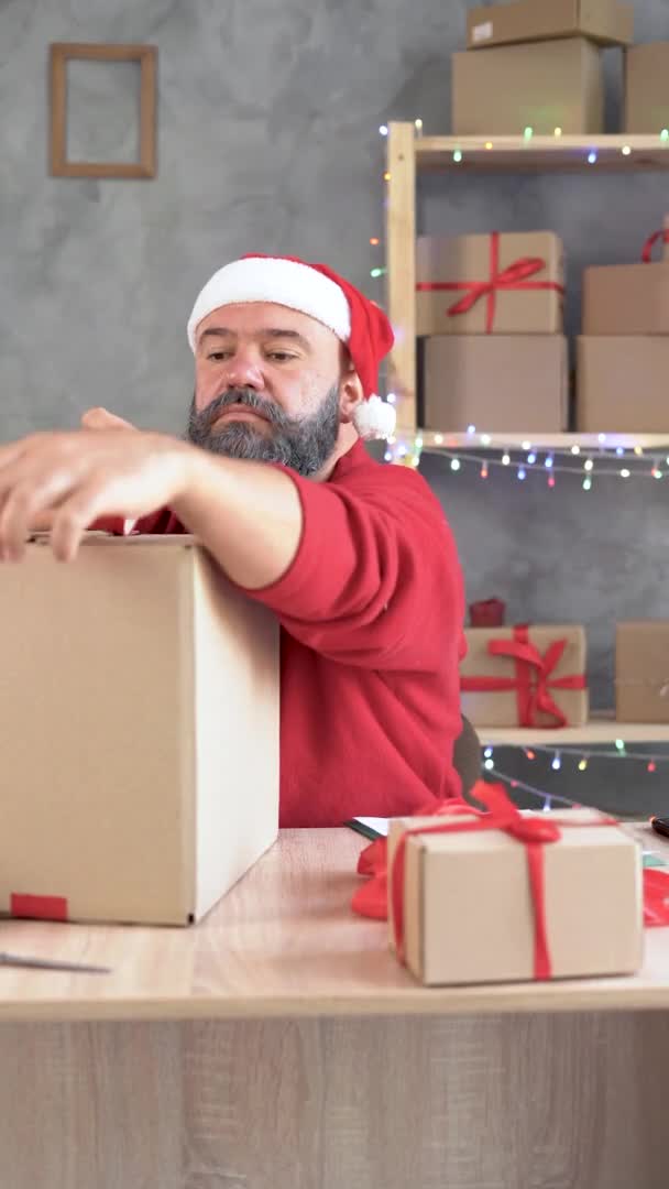 Big Christmas Sale, un homme caucasien barbu dans un pull rouge est assis à un bureau dans son bureau à la maison et enregistre une grande boîte avec des formalités administratives. prépare les marchandises pour l'expédition. — Video