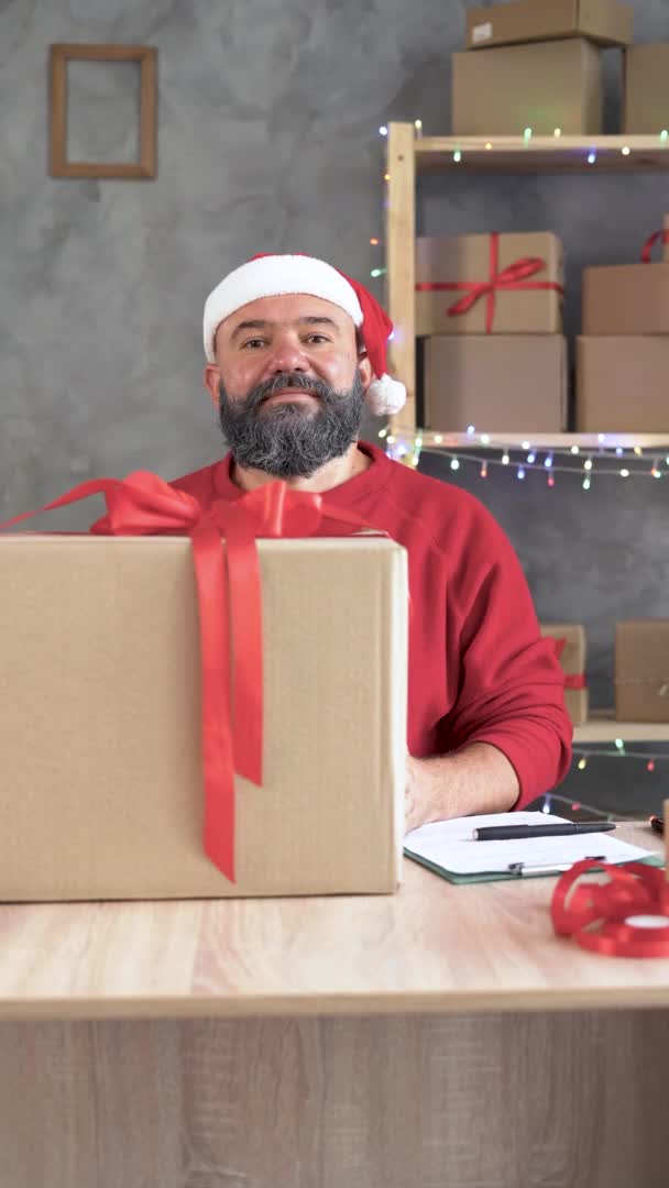 Gran venta de Navidad, un hombre caucásico barbudo en un suéter rojo se sienta en una mesa en su oficina cerca de una caja de artesanía. Sonrisa confidencial. — Vídeos de Stock