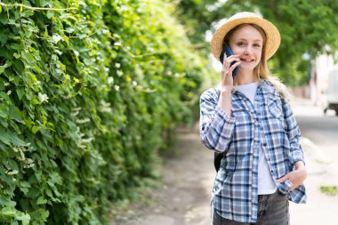 Cep telefonu kullanıcısı şapkalı kız yazın yeşil parkta dikilirken sohbet ediyor. Doğanın yeşil arka planında gezici bir turistle iletişim. yaşam tarzı konsepti