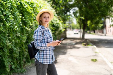 Akıllı telefonlu şapkalı ve gömlekli genç bir kadın şehirde dolaşıyor. Hippi kız internete girip cep telefonuyla kameraya bakıyor. Seyahat ve teknoloji konsepti