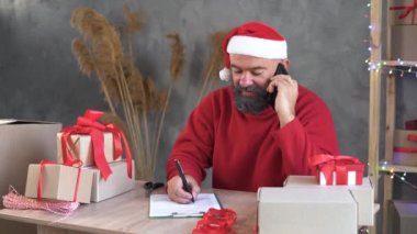 Sakallı ve Noel Baba şapkalı beyaz bir adam ev ofisinde oturuyor ve bir telefon görüşmesi için emir alıyor. Kağıda kalemle not alıyor. Yılbaşı veya yeni yıl indirimi kavramı.