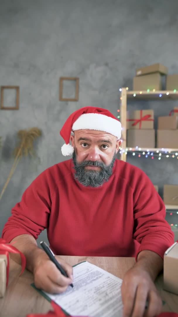 Handel online. Kaukaski mężczyzna w czapce Świętego Mikołaja i białej brodzie przyjmuje zamówienie na towary przez internet za pośrednictwem komunikacji wideo. Pionowe wideo. Koncepcja sprzedaży świątecznej na Boże Narodzenie lub Nowy Rok. — Wideo stockowe