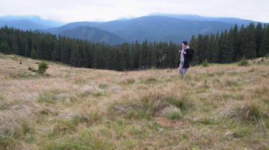 Yanlız gezgin kadın, dağ sıralarında sırt çantasıyla gezer ve şapka giyer. Rüzgar yeşil çimenleri sallıyor. Dağlarda yürüyüş kavramı.