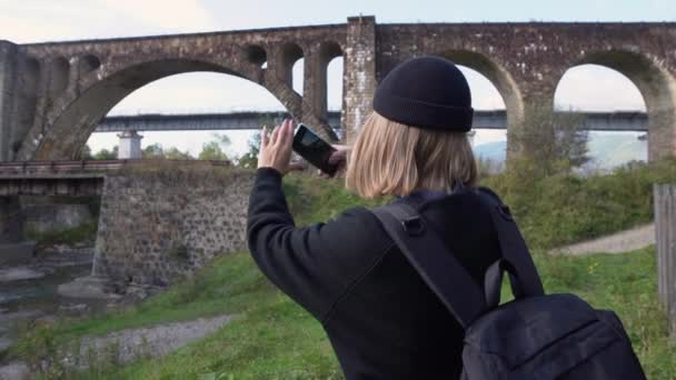 Een toerist in de herfst of voorjaar kleding neemt een foto van de historische verlaten viaduct van de spoorboog brug in Vorokhta, Ivano-Frankivsk regio, Oekraïne. — Stockvideo