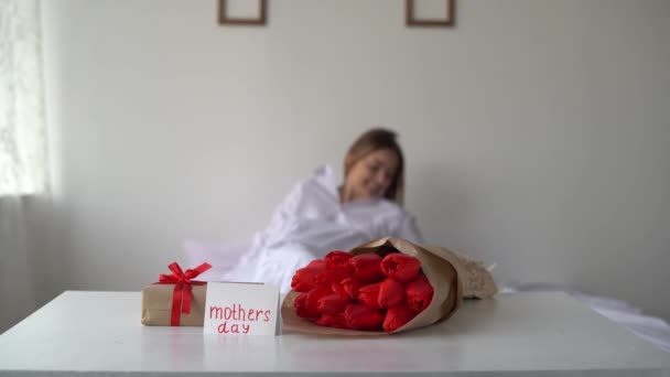 Festa della Mamma. Giovane donna al mattino in camera da letto con un mazzo di tulipani, un regalo e un biglietto di auguri per le vacanze. Concetto della festa della mamma — Video Stock