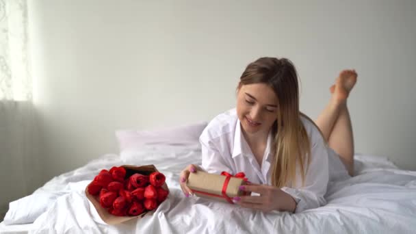 Kaukaska dziewczyna leżąca na białej pościeli z prezentem i bukietem kwiatów w jej urodziny rano w domu w sypialni korzystających relaks. — Wideo stockowe