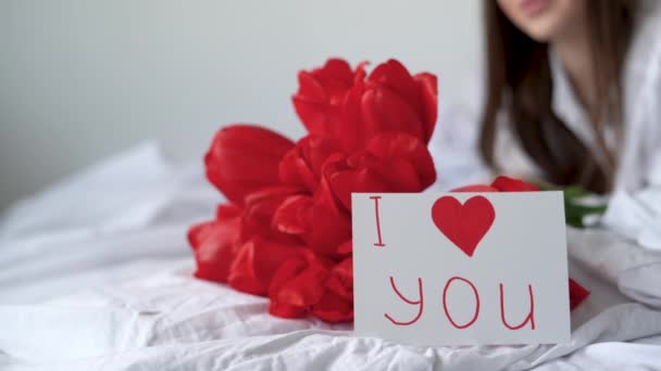 아침에 침대에 있는 소녀는 붉은 꽃 한 다발 과 발렌틴의 날이나 생일에 보내는 엽서와 함께 누워 있습니다. 생일 선물 축하 해 — 비디오