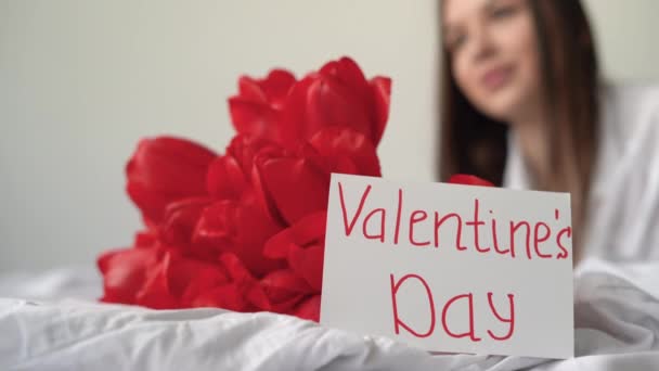 Carte cu text scris de mână Ziua Îndrăgostiților și o fată cu lalele roșii citește o carte poștală pentru vacanță dimineața în timp ce se află în pat. — Videoclip de stoc