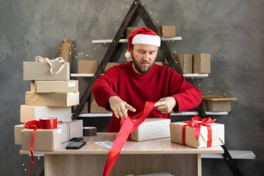 Noel Baba şapkası takan ve kırmızı süveter giyen bir erkek işadamı kutuları paketliyor ve sevkiyat için mallar hazırlıyor, kutuya kırmızı kurdele örüyor. Boşluğu kopyala.