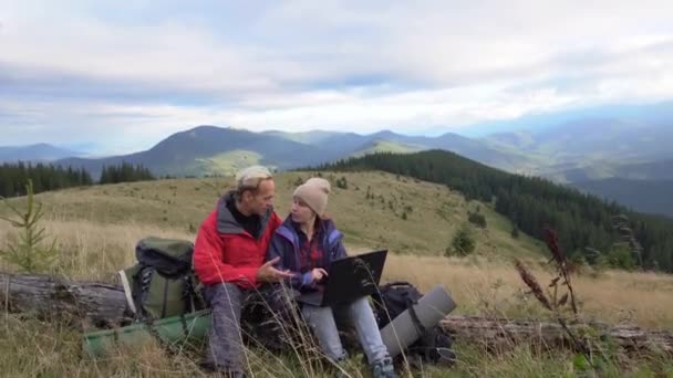 在山里度假一对男女徒步旅行者坐在一棵老树上，激烈地讨论着看笔记本电脑的问题。强风刮来了远程工作。山里的自由职业者. — 图库视频影像