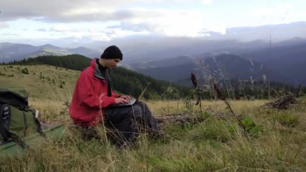 Ein erwachsener Mann arbeitet oben auf dem Berg an seinem Computer. Remote-Arbeitskonzept. Der Reisende arbeitet aus der Ferne und genießt die Berglandschaft während der Sommerferien. Videoanruf. — Stockvideo