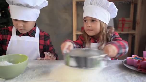 Dzieci przygotowują ciasto do pieczenia świątecznych ciasteczek w kuchni w Wigilię. Mała kucharka przesiewa mąkę przez sito w kuchni na stole. — Wideo stockowe