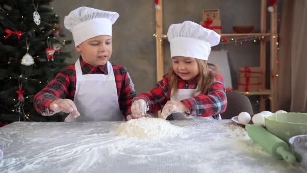 Niños divertidos en la cocina preparan masa de harina cruda para hornear galletas de jengibre de Navidad. Hermano y hermana juegan con harina vestida con el uniforme — Vídeo de stock