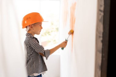 Kafkasyalı çocuk duvarları evin içinde boya rulosuyla boyuyor, kendi kendini tamir ediyor. Çocuklar, ev ya da apartman tadilatı. Turuncu kasklı çocuk, profil manzaralı. Metin için yer.