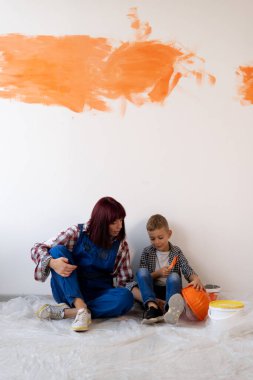 Dairede tadilat var. Mutlu aile babası ve oğlu duvarı turuncu boyayla boyarlar. Anne ve çocuk ahşap zeminde oturuyorlar. Tamirden dinleniyorlar. Ev yenileme konsepti. Boşluğu kopyala