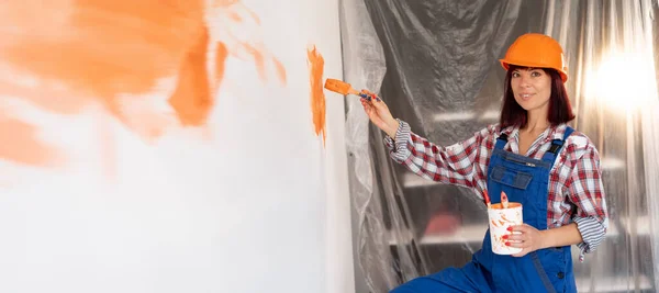 Happy caucasian woman in hard hat paints the interior wall in a new house with a brush in orange. Construction and renovation concept. Home renovation. Background and place for text. Banner.