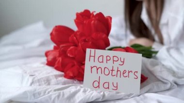 Anneler Günün kutlu olsun. Yatakta uzun saçlı bir kız, elinde bir buket kırmızı lale ve üzerinde yazı olan bir kartpostal var. Bir kadına çiçekler.