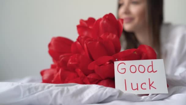 A girl wakes up in the morning in the bedroom on white bedding and sees a bouquet of red tulips on the table with a postcard with the good luck. — Stock Video