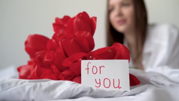 Chica en la cama con un ramo de tulipanes rojos y una postal con una inscripción para usted. Flores para una mujer para su cumpleaños o San Valentín. — Vídeo de stock