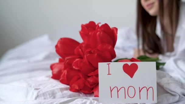 Feliz Dia das Mães. Uma jovem acorda de manhã no quarto e vê um buquê de flores um presente e um cartão de felicitações. tulipas de uma criança — Vídeo de Stock